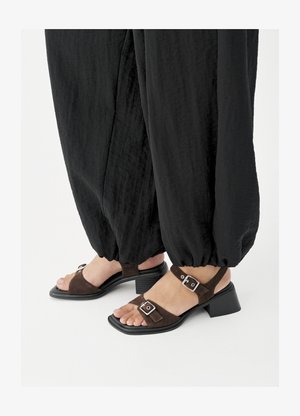 Kājām iespējotas brūnas zamšādas sandalītes ar plata papēža bloku un sudraba sprādzēm, kopā ar vaļīgām melnām biksēm ar aprocēm uz baltas virsmas.