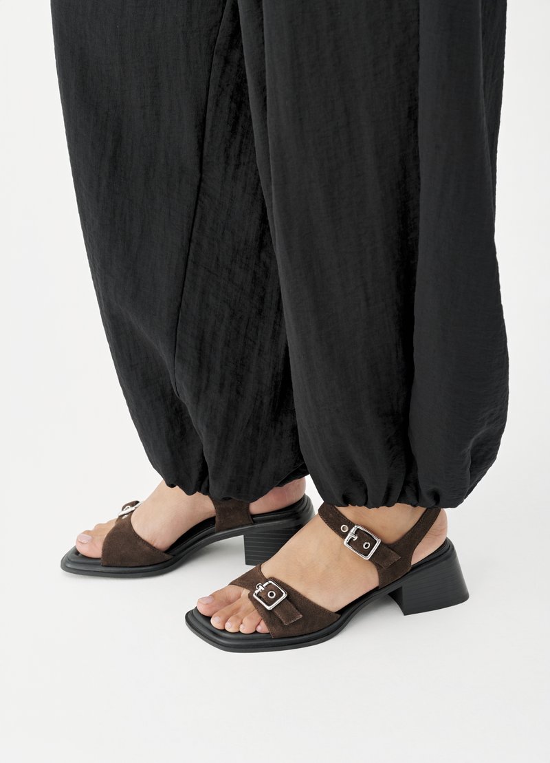 Füße in braunen Wildleder-Sandalen mit Blockabsatz und silbernen Schnallen, kombiniert mit lockeren schwarzen, umgeschlagenen Hosen auf weißem Hintergrund.