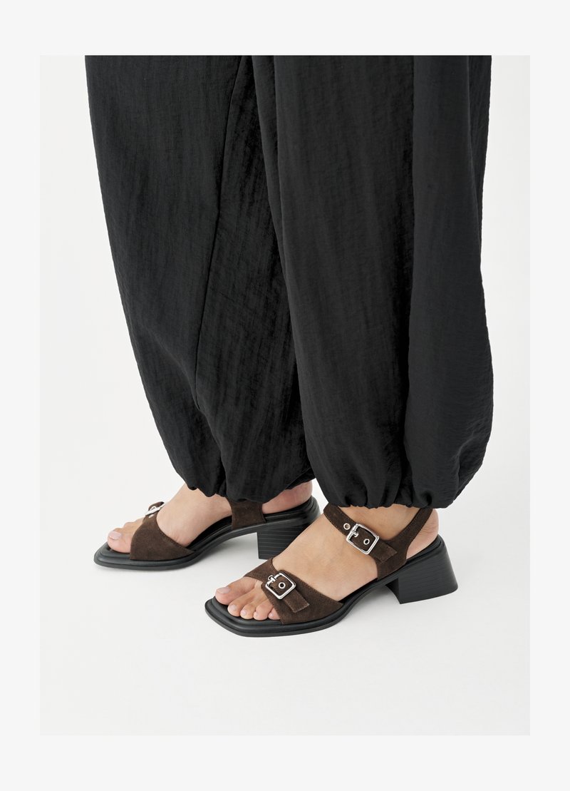 Füße in braunen Wildleder-Sandalen mit Blockabsatz und silbernen Schnallen, kombiniert mit lockeren schwarzen, umgeschlagenen Hosen auf weißem Hintergrund.