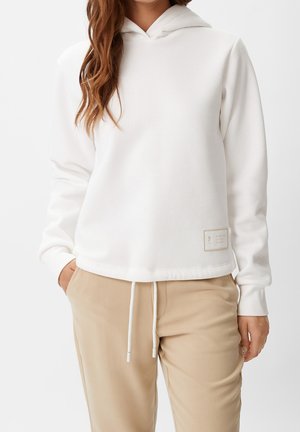Frau trägt weißen Kapuzenpullover und beige Kordelzughose, mit einer Hand in der Tasche, gezeigt vom Hals bis zur Hüfte.