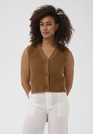 Femme aux cheveux bouclés portant un gilet en tricot marron sans manches boutonné et un pantalon blanc, debout devant un fond uni.