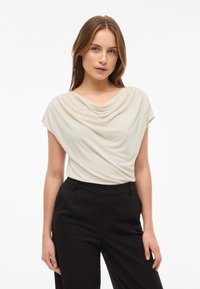 Kvinne med lys brunt hår iført en kremfarget drapert utenermet topp og sorte høyhvitde bukser foran en enkel hvit bakgrunn.