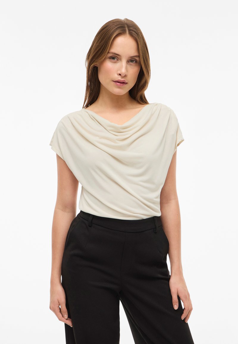 Kvinne med lys brunt hår iført en kremfarget drapert utenermet topp og sorte høyhvitde bukser foran en enkel hvit bakgrunn.