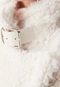 Textil blanco esponjoso con una textura gruesa, que presenta un cinturón de cuero claro con una hebilla plateada y detalles de costura.