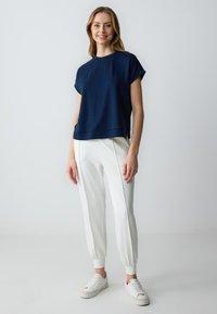 Top navy a maniche corte con una vestibilità rilassata, abbinato a pantaloni bianchi a sigaretta e sneaker bianchi, il tutto su uno sfondo leggero e uniforme.