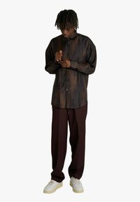 Camicia marrone con fantasia, chiusura con bottoni e taglio comodo, abbinate a pantaloni larghi color bordeaux e sneakers bianche, che mostrano una superficie testurizzata.