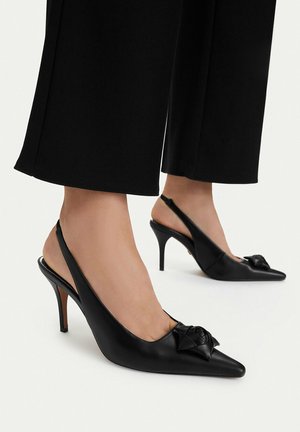 Czarne czółenka typu slingback z szpiczastym noskiem i ozdobną kokardą, noszone z czarnymi, krótkimi spodniami na jasnym tle.