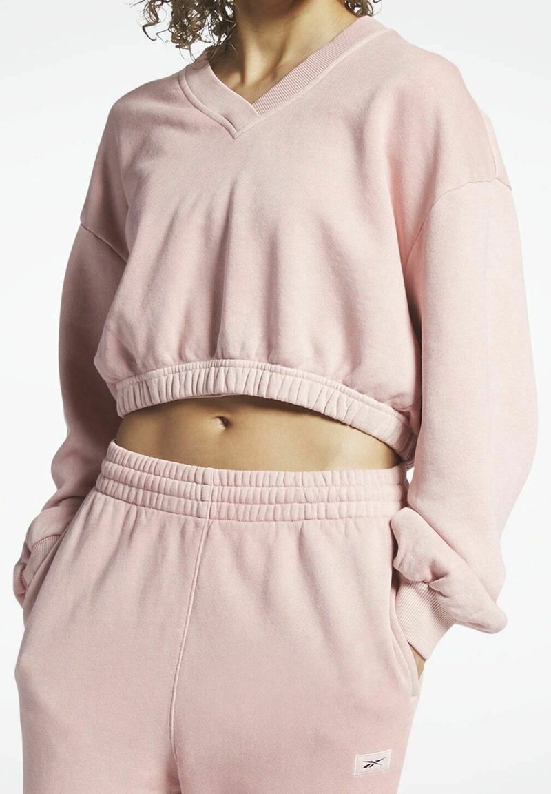 Femme portant un sweat-shirt court rose clair et un pantalon de survêtement taille haute assorti, mains dans les poches, montrant le ventre.