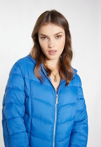 Jachetă puffer albastră cu fermoar frontal, textură matlasată și glugă. Modelul poartă un colier cu pandantiv auriu și are părul coafat lejer.