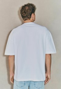 Camiseta de algodón blanca de manga corta con un corte relajado, cuello redondeado y una textura suave. Lleva pantalones de mezclilla azul claro.
