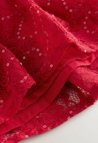 Czerwony koronkowy materiał z kwiatowym wzorem i cekinami, nałożony na jednolity czerwony materiał, ukazujący bogatą teksturę i detale.