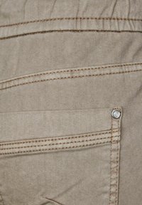 Tejido beige con una textura suave, que presenta costuras marrones contrastantes y un detalle de herrajes plateados en un bolsillo trasero.