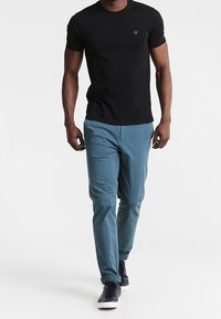 T-shirt nera a maniche corte con un piccolo logo sul petto, abbinata a pantaloni blu chiaro e sneaker scure, mentre cammina su uno sfondo neutro.