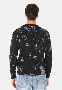 Maglione nero a maglia con motivo splatter bianco, scollo rotondo, polsini e orlo a costine, e vestibilità rilassata, indossato sopra jeans azzurri chiari.