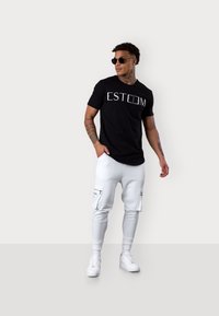 Schwarzes T-Shirt mit weißem "ESTEEM"-Text, kombiniert mit weißen Jogginghosen mit seitlichen Reißverschlusstaschen und schmalen Bündchen, getragen mit schwarzen Sonnenbrillen.