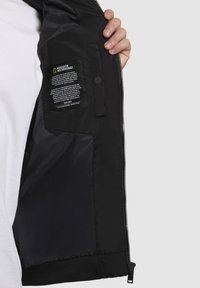 Schwarze isolierte Jacke mit einem glänzenden Innenfutter, einem Reißverschluss vorne, geripptem Saum und einem Etikett mit Pflegehinweisen auf der linken Seite.