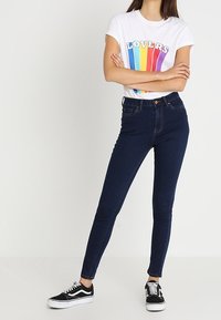 Camiseta blanca con un gráfico colorido de "LOVERS", combinada con jeans ajustados de color azul oscuro y zapatillas negras con suelas blancas.
