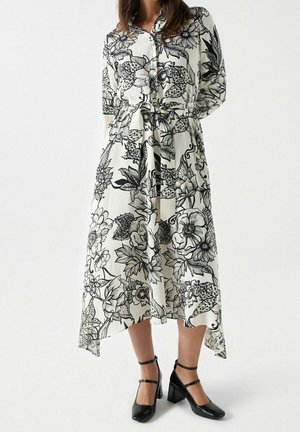 Kobieta w białej sukience midi z czarnym kwiatowym wzorem, z paskiem w talii, z długimi rękawami i czarnych butach na słupku z paskiem przy kostce.