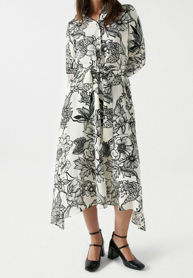 Femme portant une robe midi blanche à imprimé floral noir, taille ceinturée, manches longues et chaussures à talons blocs noires avec bride à la cheville.