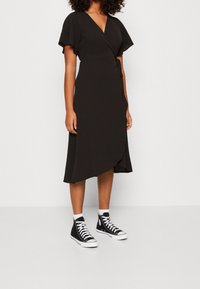 Černé zavinovací šaty s krátkými rukávy, výstřihem do V a detailem na bočním zavázání; doplněné černými vysokými teniskami a bílými ponožkami. Hladká textilie.