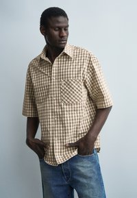 Hombre con camisa de manga corta beige a cuadros y vaqueros azules, de pie con las manos en los bolsillos, mirando hacia abajo a su izquierda.
