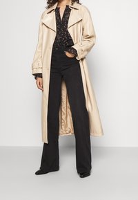 Beige trenchcoat med brede revers, lange ærmer og knaplukning ved manchetterne; parret med en sort mønstret bluse og højtaljede sorte jeans.