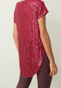 Top de lentejuelas rojas con mangas cortas, dobladillo redondeado y un acabado texturizado; llevado sobre pantalones ajustados burdeos.
