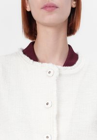 Biała kurtka o teksturowanym wykończeniu z okrągłym kołnierzem i bordowym podszyciem, z pięcioma guzikami z złotymi akcentami z przodu.