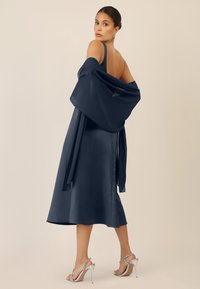 Abito in satin navy con una silhouette fluida e una schiena bassa, abbinato a uno scialle coordinato. Sandali alti argentati completano il look.