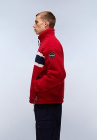 Jachetă din fleece roșu cu guler înalt, prezentând dungi albastre și albe pe piept și o etichetă cu logo rotund pe mâneca stângă.