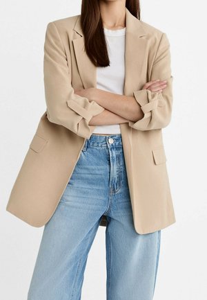 Kvinde med korslagte arme iført en beige blazer over en hvid top og lyseblå jeans med høj talje mod en ensfarvet baggrund.