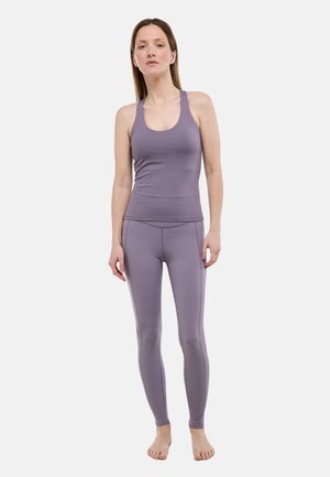 Femme debout pieds nus, portant un débardeur mauve ajusté à dos nageur et un legging taille haute assorti, sur un fond uni.