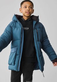 Veste matelassée teal avec capuche, dotée d'une fermeture éclair à l'avant, de poches latérales et d'accents noirs ; portée par-dessus un sweat à capuche noir.