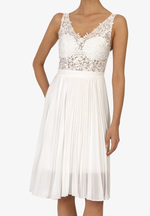 Robe blanche avec un corsage en dentelle à motifs floraux et une jupe plissée. La robe possède un décolleté en V et est sans manches.