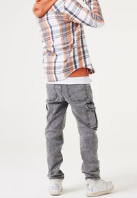 Persona che indossa pantaloni cargo grigi, sneaker bianche Nike, felpa arancione e camicia a quadri, in piedi contro uno sfondo chiaro e semplice.