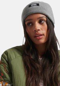 Bonnet tricoté gris avec une étiquette noire à l'avant. Porté avec une veste bombardier vert olive ornée de broderies colorées.