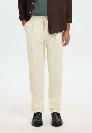 Homme portant un pantalon crème avec taille à cordon, une chemise marron foncé déboutonnée, un sous-pull vert et des chaussures en cuir noir.