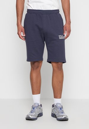 Mann trägt marineblaue elastische Shorts mit weißem Text am linken Oberschenkel, weiße Sportsocken und graue Sportschuhe auf weißem Boden.