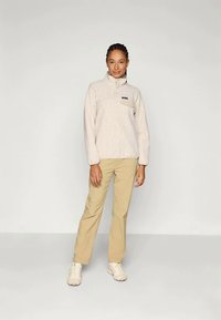 Žena stojaca v béžovom fleecovom pulóvri, svetlohnedých nohaviciach a bielych teniskách na jednoduchom svetlom pozadí.