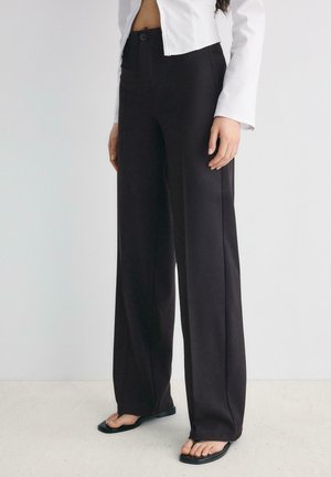 Personne portant un pantalon noir taille haute à jambes larges, une veste blanche courte et des tongs noires à bout carré, debout sur un sol clair.