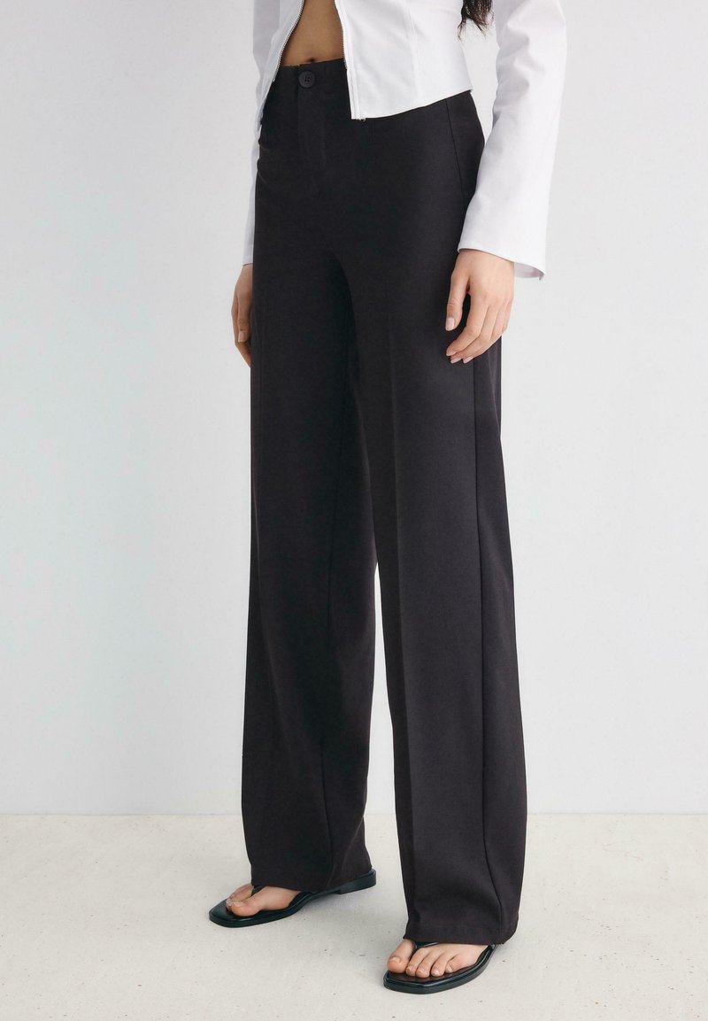 Personne portant un pantalon noir taille haute à jambes larges, une veste blanche courte et des tongs noires à bout carré, debout sur un sol clair.