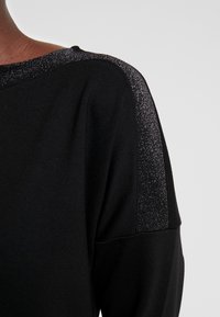 Sweat-shirt noir avec une texture douce, agrémenté d'accents argentés scintillants sur la couture de l'épaule. Design décontracté avec une coupe ample.