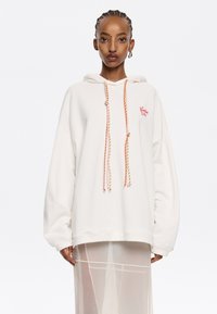 Sudadera blanca de gran tamaño con cordones en naranja y crema, que presenta un pequeño logo rojo en el pecho y un tejido suave y texturizado.