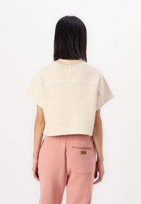 Felpa cropped beige con scollatura rotonda, orli grezzi e maniche corte, abbinata a pantaloni della tuta rosa con tasca posteriore e logo.