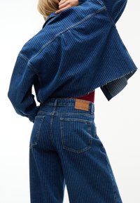 Άτομο που φοράει ταιριαστό μπλε ριγέ κοντό denim μπουφάν και ψηλόμεσα τζιν με καφέ δερμάτινη ετικέτα "Tommy Jeans" στη μέση.