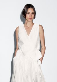 Kobieta z krótkimi ciemnymi włosami, ubrana w bezrękawnik w kolorze białym i dopasowaną spódnicę, stoi z rękami w kieszeniach na jednolitym tle.