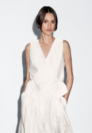 Kvinna med kort mörkt hår bär en ärmlös vit wrap-top och matchande kjol, står med händerna i fickorna mot en enkel bakgrund.