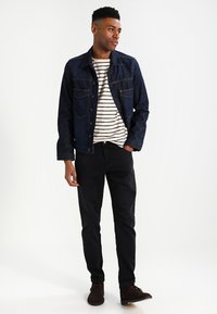 Donkerblauwe denimjack met knoopsluiting en borstzakken, gecombineerd met een wit en navy gestreept t-shirt en zwarte broek, afgerond met bruine schoenen.