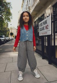 Chaleco de mezclilla con botones, camiseta roja de manga larga, pantalones de cuadros en blanco y negro, y zapatillas plateadas. De pie en una acera de la ciudad.