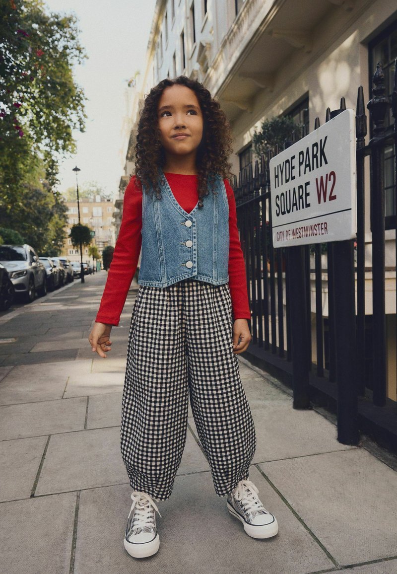 Chaleco de mezclilla con botones, camiseta roja de manga larga, pantalones de cuadros en blanco y negro, y zapatillas plateadas. De pie en una acera de la ciudad.